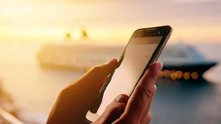 Mano sosteniendo un móvil al atardecer con un crucero de fondo, consultando datos móviles para viajar en el extranjero