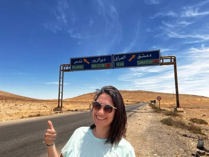 Cris sonriendo con gafas de sol y el pulgar hacia arriba bajo un cartel de carretera en árabe, en pleno desierto