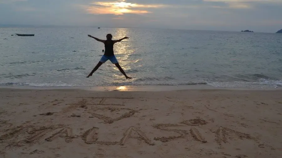 Persona saltando con los brazos abiertos en una playa al amanecer, con una palabra escrita en la arena