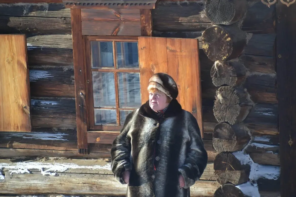 Mujer con abrigo de piel junto a una casa de madera tradicional siberiana