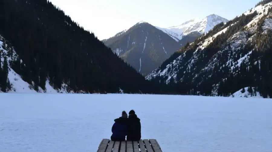 Dos viajeros sentados en un banco frente al lago Kolsai helado y montañas nevadas