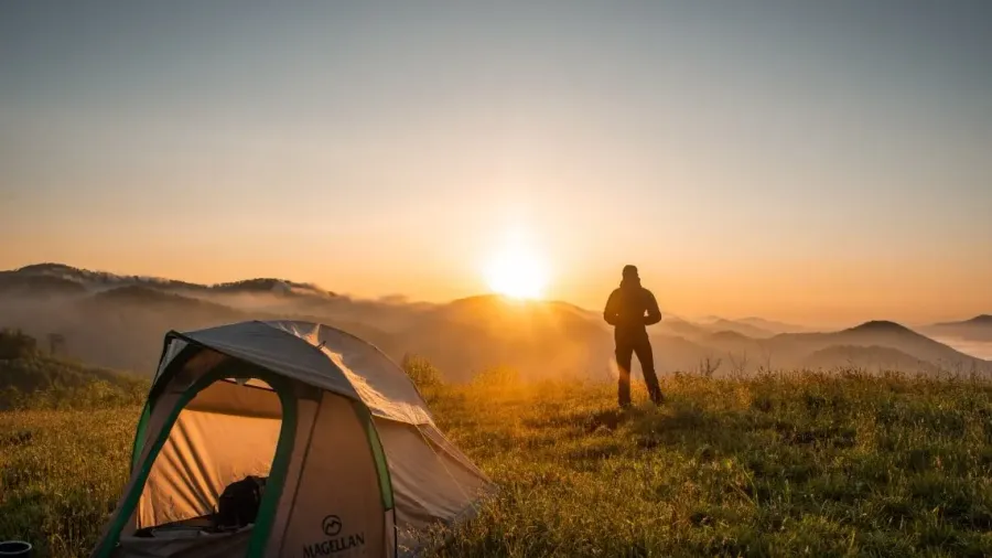 Persona de pie frente a una tienda de campaña al amanecer sobre un paisaje de montañas