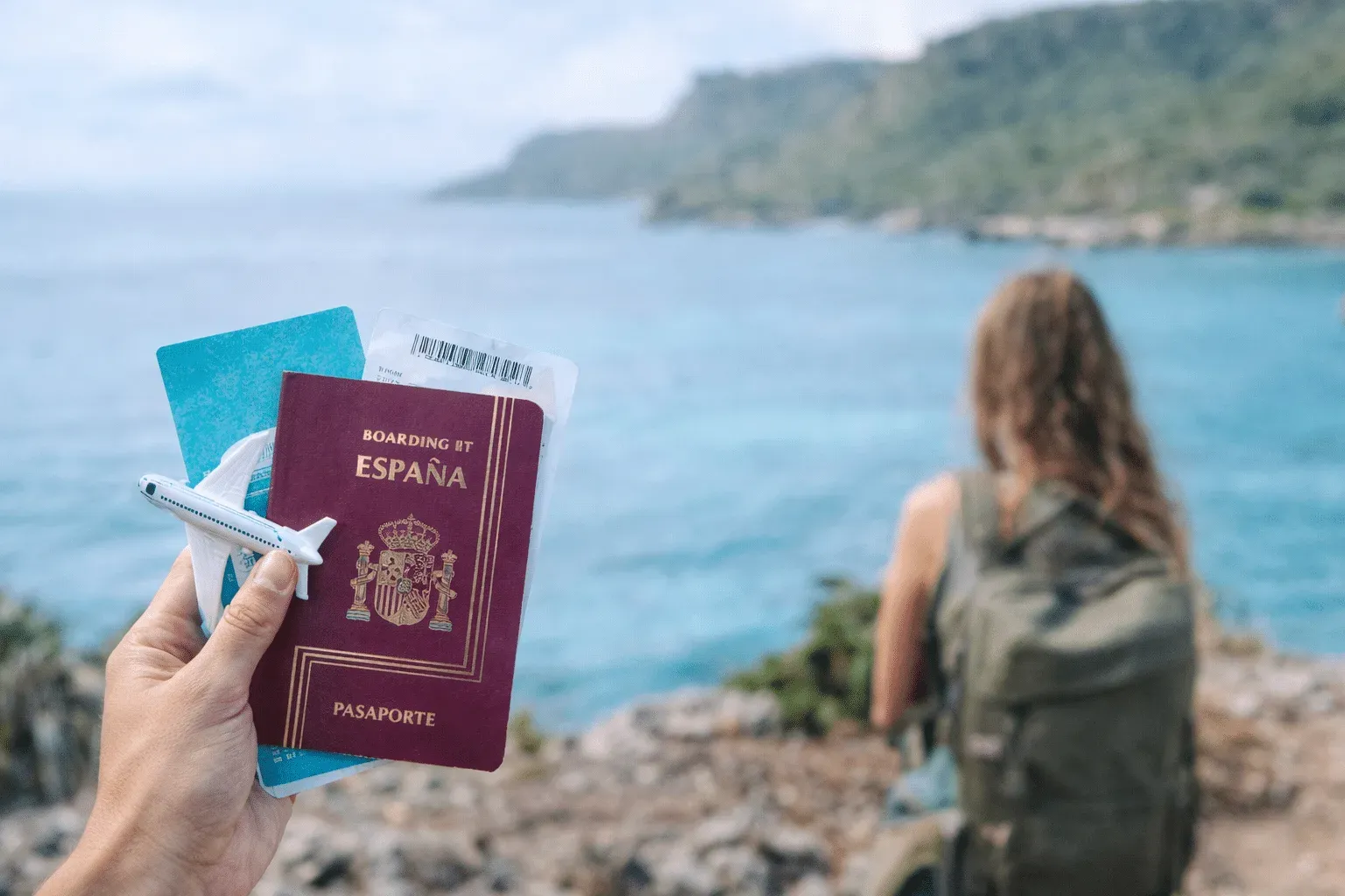 Mano sosteniendo un pasaporte español, tarjetas de embarque y un avión de juguete frente al mar, con una viajera con mochila de espaldas