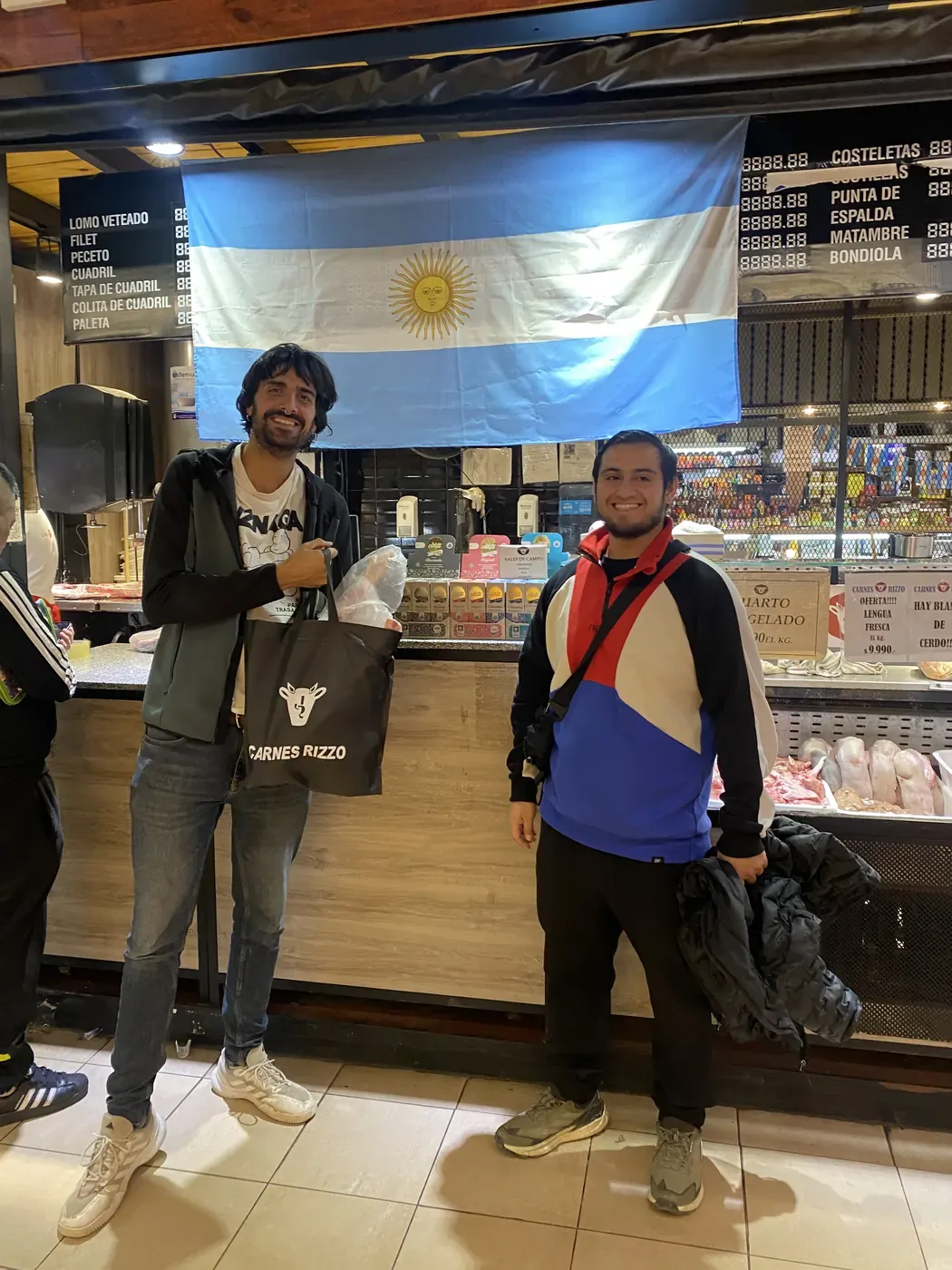 Dos amigos posan frente a una carnicería con una bandera argentina; uno sostiene una bolsa de Carnes Rizzo
