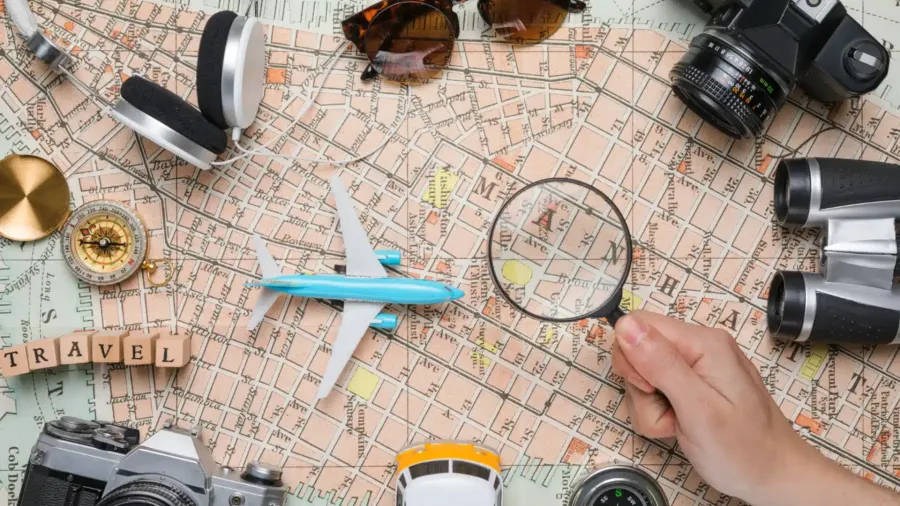 Mapa antiguo de una ciudad con una mano sosteniendo una lupa, un avión de juguete azul, una brújula, unos prismáticos, gafas de sol, auriculares, dos cámaras de fotos y la palabra TRAVEL formada con cubos de madera