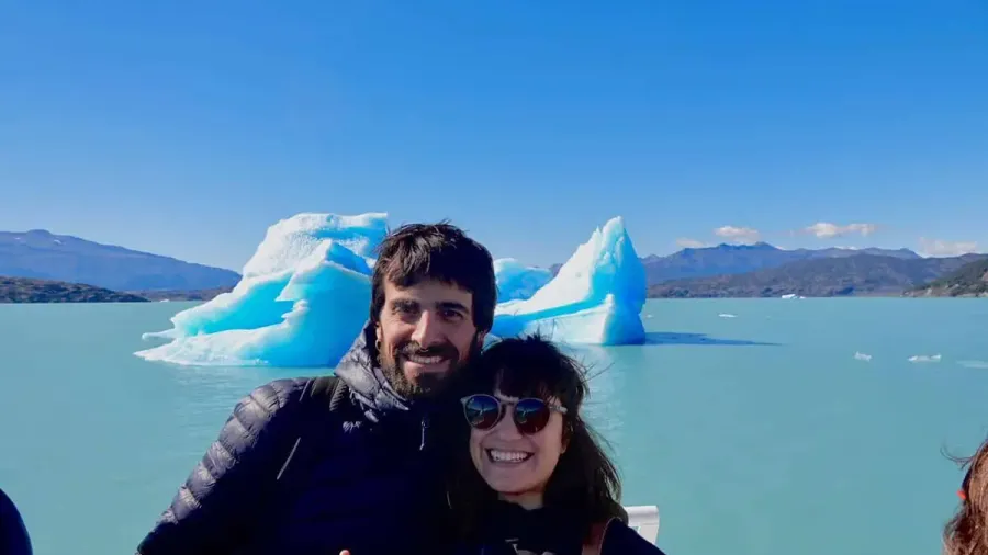 Sergio y Cris sonriendo a bordo de un barco en el Lago Argentino, con un témpano de hielo azul de fondo