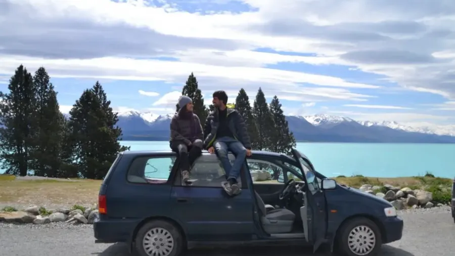 Dos viajeros sentados sobre un coche junto a un lago turquesa con montañas nevadas