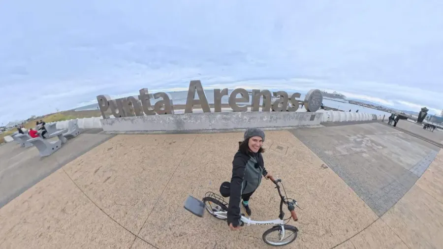 Cris en bicicleta plegable frente al cartel de letras gigantes de Punta Arenas, junto al Estrecho de Magallanes