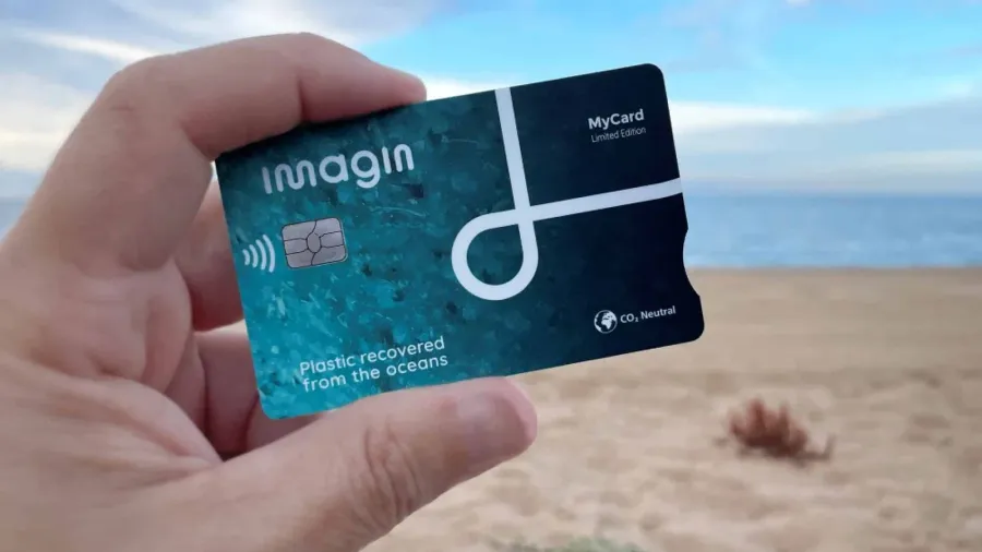 Mano sosteniendo una tarjeta imagin azul petróleo, edición limitada hecha con plástico recuperado de los océanos, frente a una playa con el mar y un cielo con nubes de fondo
