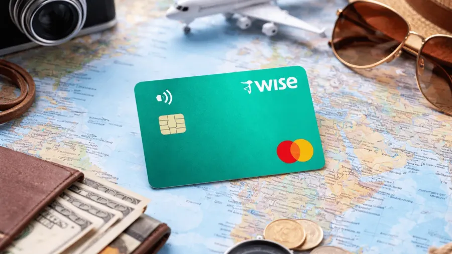 Tarjeta Wise verde con logo Mastercard sobre un mapamundi, rodeada de una cámara de fotos, un avión de juguete, gafas de sol, un sombrero de paja, una brújula, monedas y una cartera con billetes de dólar